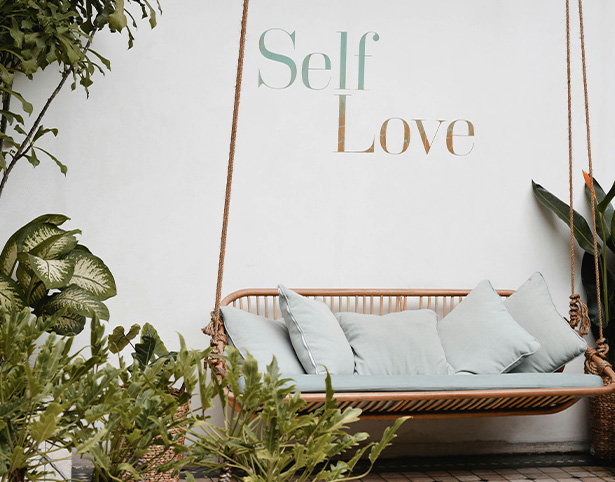 Hammock with cushions in front of a wall displaying the text Self Love, surrounded by greenery.