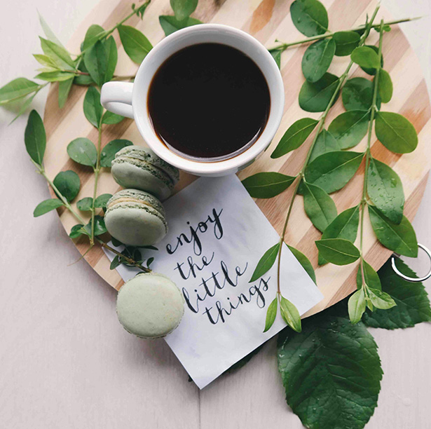 A cup of coffee with green macarons on a wooden platter surrounded by leaves.